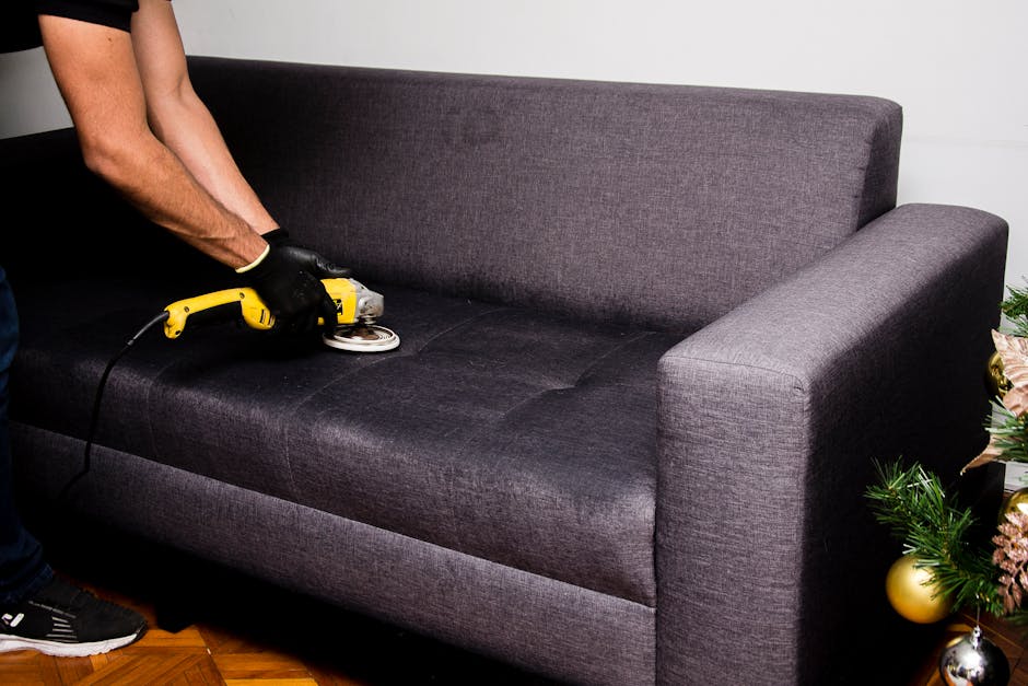 A person in a black jacket is using a handheld vacuum cleaner to perform surface cleaning on a decorative yellow and white patterned cushion placed on a dark fabric sofa. The sofa is situated against 