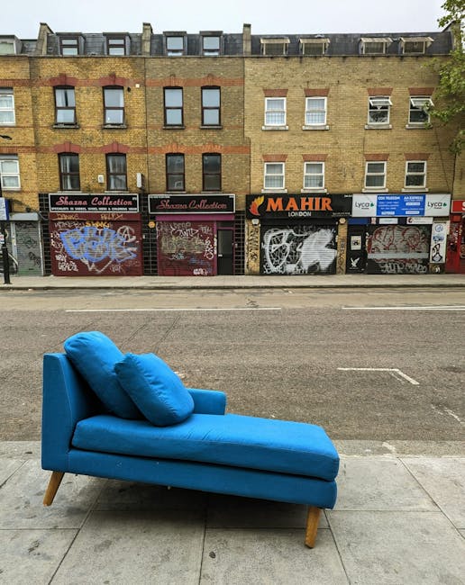 A bright blue upholstered chaise lounge with two matching cushions is positioned outdoors on a sidewalk in front of a row of commercial storefronts with closed shutter doors covered in graffiti. The f