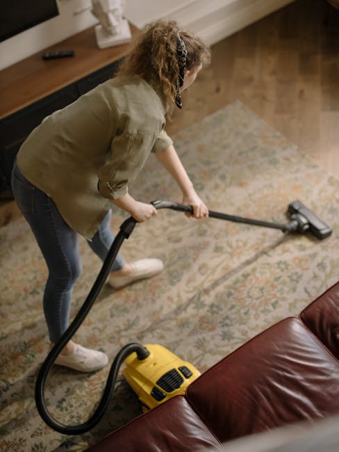 Close-up of a vacuum cleaner head actively cleaning a patterned area rug with intricate floral motifs, situated on a wooden floor in a well-lit room. The vacuum cleaner is black and modern, with a sle
