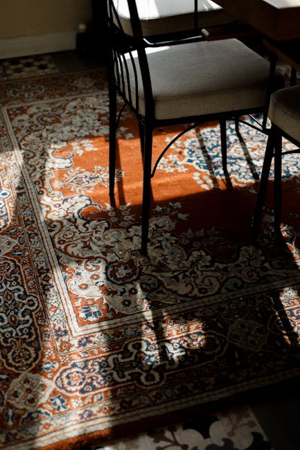 A woman in casual home attire, including a yellow top and light-colored pants, is vacuuming a dark grey carpet in a bright, modern living room. The room features large windows with natural daylight, s